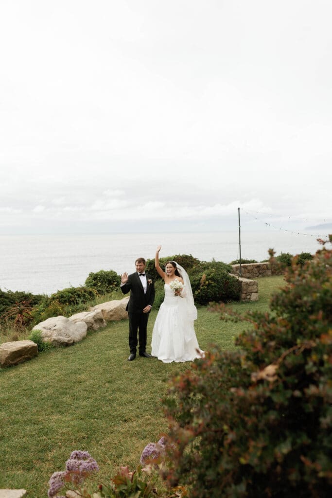 Newlyweds waving to guests as they walk across the cliffside lawn after their ceremony.