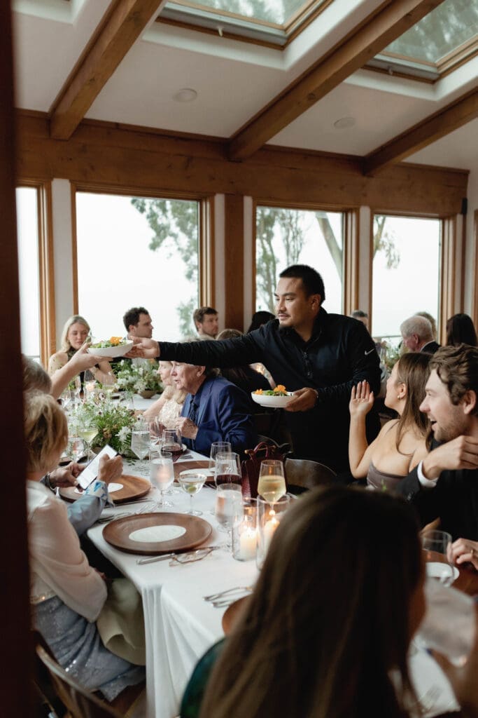 Private caterer serving plates to laughing guests seated at the reception table.