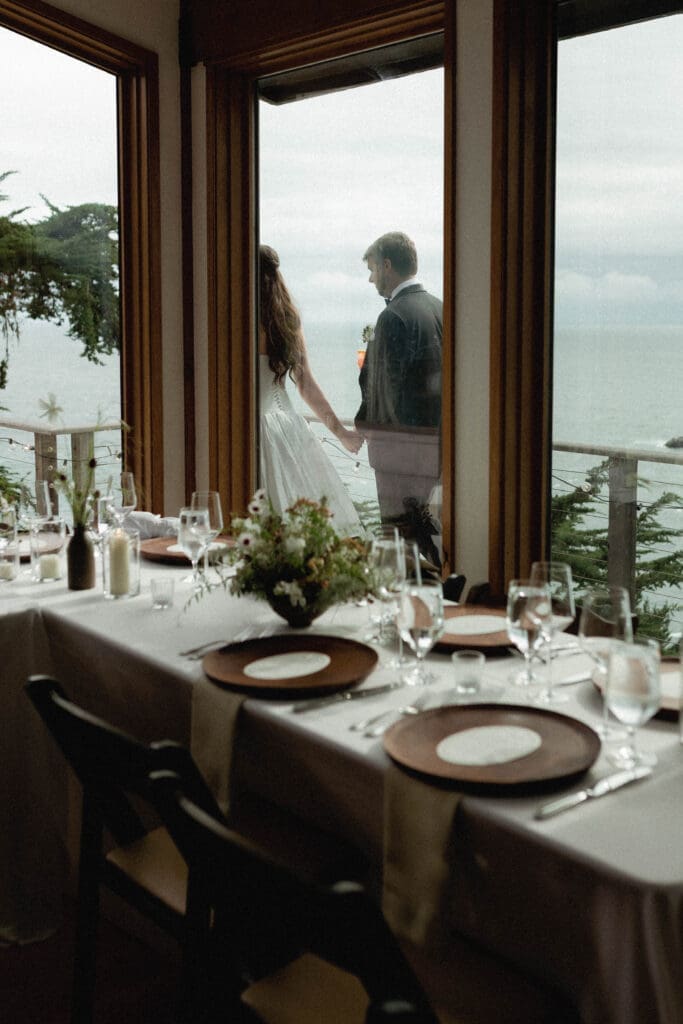 Bride and groom visible through the floor-to-ceiling windows of the reception space at Wind and Sea Estate, reflected over the set dinner table.